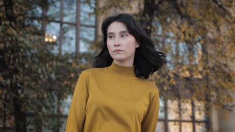 Young girl in mustard yellow colour sweatshirt standing outside, during windy weather. Beautiful asian young woman waiting alone on background of trees and building, gimbal shot