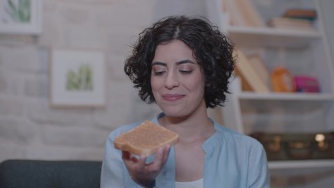 Young woman is sitting on an armchair at home and eating peanut butter bread.