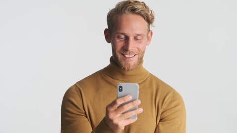 Attractive positive blond bearded man intently using smartphone and smiling over white background