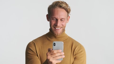 Young attractive blond bearded man using smartphone and rejoicing on camera over white background. Yes gesture. Win concept