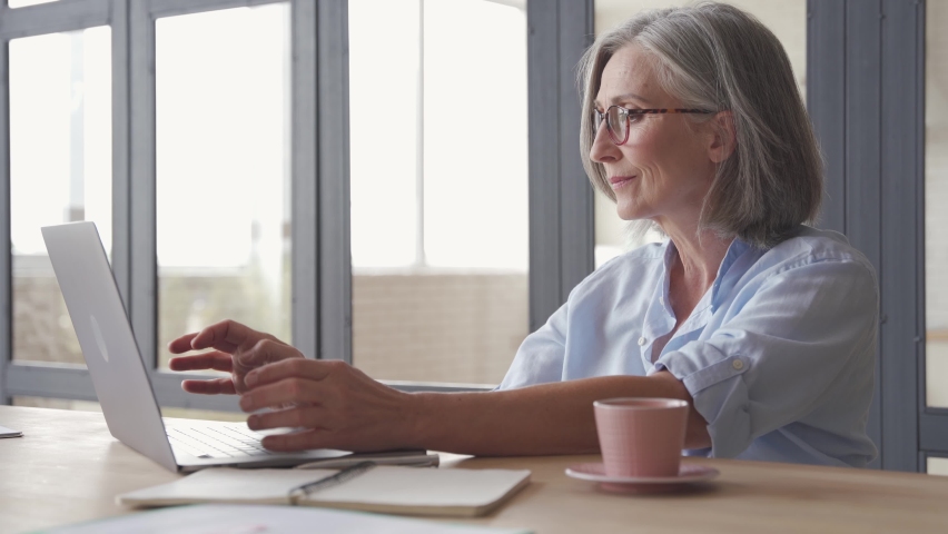 Older professional mature business woman using laptop computer sitting at workplace desk. Happy senior older employee 60s grey-haired businesswoman executive working typing on pc at home from office Royalty-Free Stock Footage #1061284702