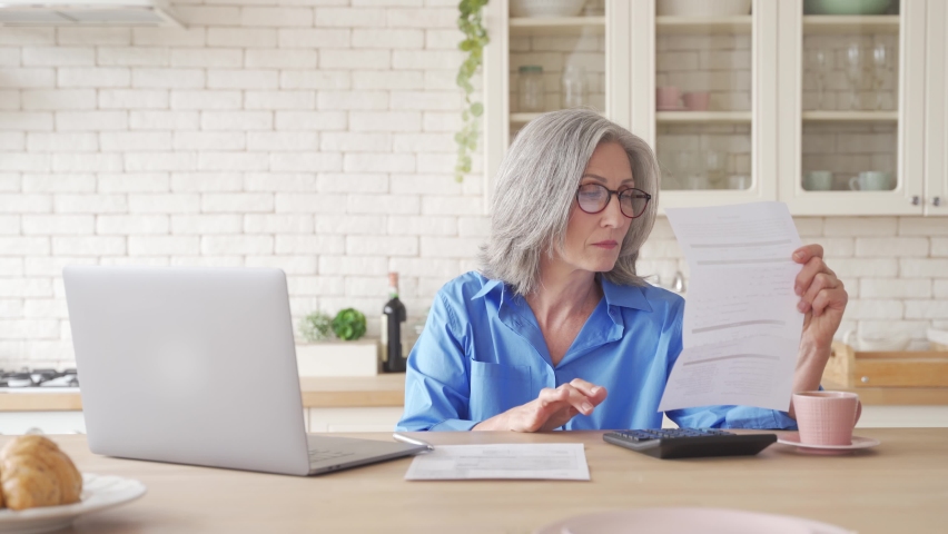 Serious worried old senior woman reading paper bill pay online at home, managing bank finances, calculating taxes, fees, high rental rates, planning loan debt pension payment sit at kitchen table. Royalty-Free Stock Footage #1061284705