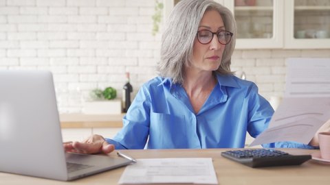 Serious worried old senior woman reading paper bill pay online at home, managing bank finances, calculating taxes, fees, high rental rates, planning loan debt pension payment sit at kitchen table.