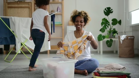 african woman folding clean clothing from clothes dryer in basket Spbd. female mother do domestic chores, housekeeping laundry while happy little afro daughter is having fun on background.