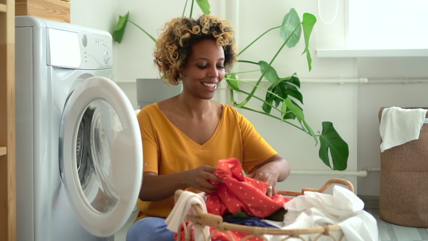 Beautiful young woman putting clothes into washing machine, sitting on floor in laundry room spbd. African American female puts dirty clothing inside of household appliance and smiles, sits in bright Royalty-Free Stock Footage #1061285125