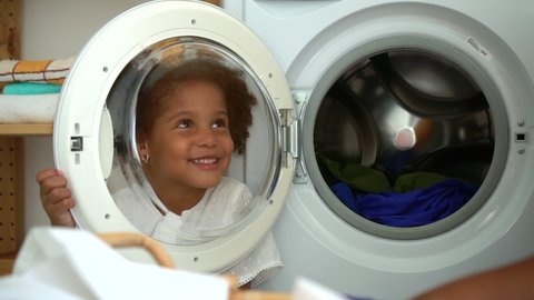 Young American mom and cute daughter having fun together in laundry room at home spbd. Cheerful woman and little girl have good time in bright interior, child looks through door of washing machine and