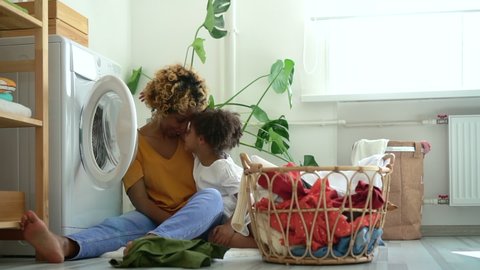 Young mother and little daughter hug and sit on floor in laundry room at home spbd. African American woman and cute girl hugging and having fun with smiles, sitting in bright interior with washing