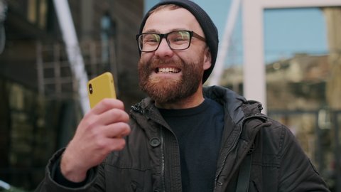 Close up of guy in glasses looking at phone screen and rejoicing while walking at street. Bearded male person using smartphone and having good news. Concept of success and victory
