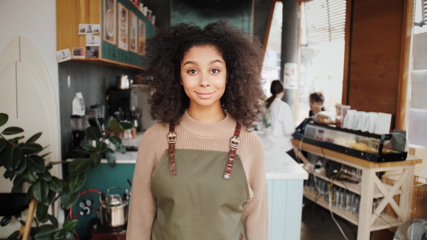 Happy stylish African American woman, generation z female with Afro hair looks at camera stands in cafe interior. Smiling mixed race latin young woman headshot portrait. Work for international Royalty-Free Stock Footage #1061289886