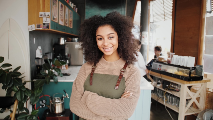 Happy stylish African American woman, generation z female with Afro hair looks at camera stands in cafe interior. Smiling mixed race latin young woman headshot portrait. Work for international Royalty-Free Stock Footage #1061289886