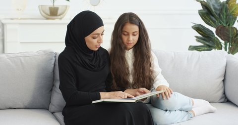 Young Arabic woman in black headscarf reading interesting story in book for pretty small teen girl. At home on sofa. Muslim mother learning and educating little teenage daughter with textbook.