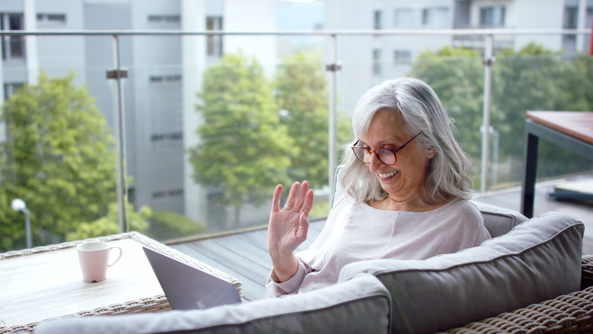 Senior woman with laptop on balcony at home, video call with family.