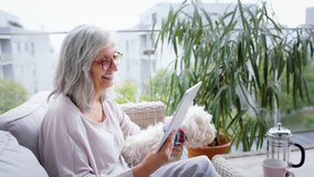 Senior woman with dog and tablet on balcony at home, video call with family.