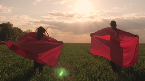 happy girls play superheroes they run across green field in red cloak, cloak flutters in wind. children's games and dreams. Slow motion. teenager dreams of becoming superhero. young girls in red cloak