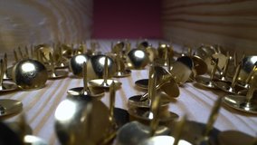Camera moves over the scattered pushpins. Super macro shot of golden shiny office supplies that lie in a wooden box. Vertical slide back.