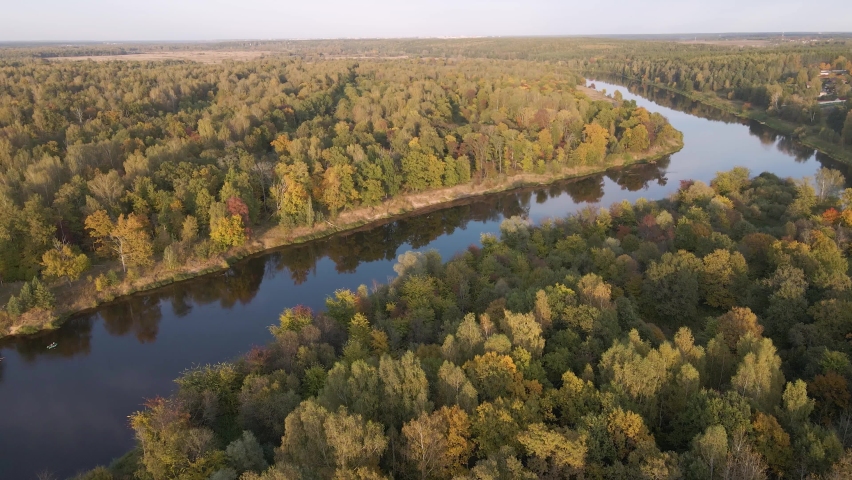 Flight in the evening over the river with densely overgrown banks. The leaves of the trees are colored yellow, orange and red. In the distance the sky at sunset. Autumn season. Soft light. 4K
