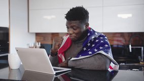 African american man with USA flag over his shoulders watching game on his laptop with cup of tea on the table. Surprised wace expression. High quality 4k footage - Powered by Shutterstock - Get 15% off with code: PIKWIZARD15