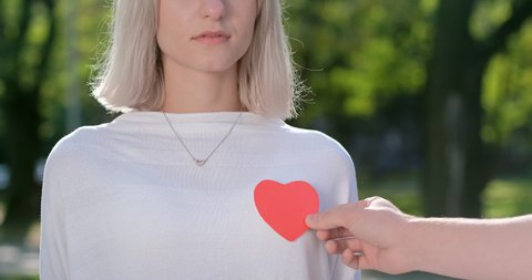 A guy with his hand clings the red paper heart to a girl in a white sweater in the area of a real heart. Give a heart to a loved one.