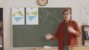 Young Caucasian female teacher writing on equation on chalkboard and explaining it at camera while giving online lesson from school during coronavirus quarantine