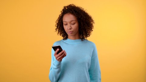 Good luck and winnings. young beautiful dark-skinned woman looks at the smartphone with great hope crossing her fingers. woman jumping for joy. in a blue sweater. person on isolated yellow background