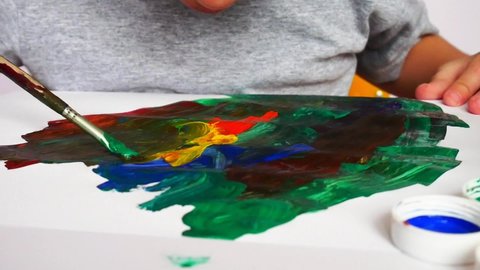 A little boy smears gouache with a brush on the sheet of paper learning to paint