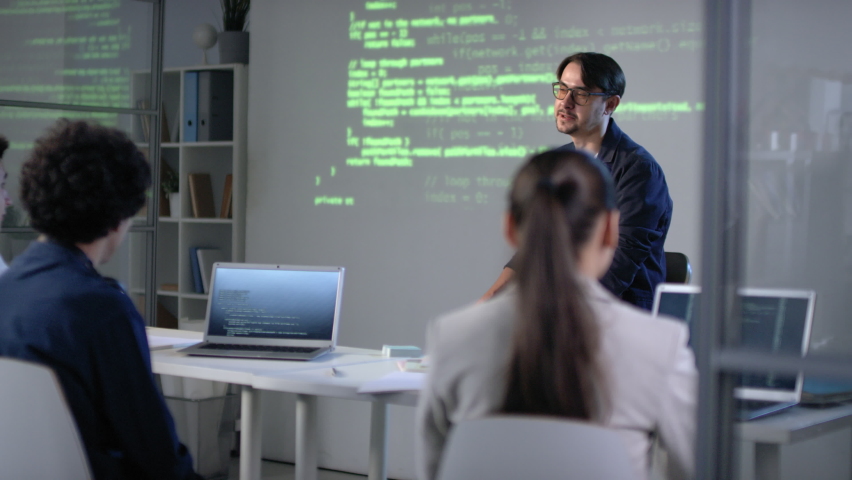 Medium shot of information technologies teacher sitting in front of teenage students with laptops giving lecture about coding programs referring to screen projector | Shutterstock HD Video #1061315044
