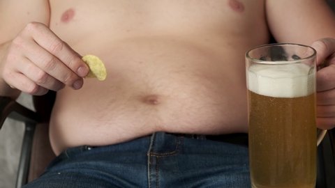 The fat man is eating chips and drinking beer. The hand scratches the beer belly. Beer belly from harmful food. Bad food, gluttony. Close-up