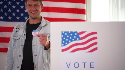 Young smiling American holds a vote sticker in front of a voting booth. The man is wearing a denim jacket. US elections concept