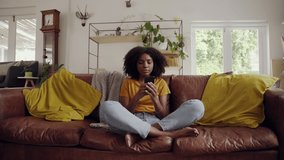Smiling relaxed afro american woman sitting on sofa hold smart phone watching social media video in living room