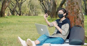 Young woman in protective mask recording video or having video communication using smartphone sitting on the grass in the park. Distance work, education and blogging concept