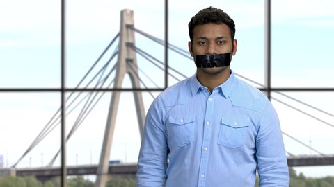 Portrait of Indian man with black tape over mouth. Cables and tower of the suspension bridge on the background. Censorship concept.