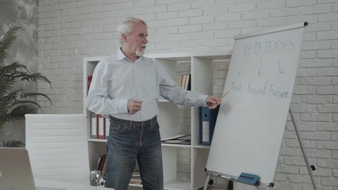 Smart mature tutor conducts language classes online. Intelligence gray haired male teacher points on whiteboard, a laptop on the table in front. Online education concept