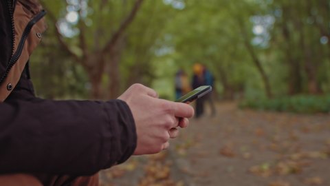 Young guy with a phone in his hands sitting on a bench in the park. In the background, a group of friends walking in the park. The concept of loneliness and dependence on the Internet and smartphones.