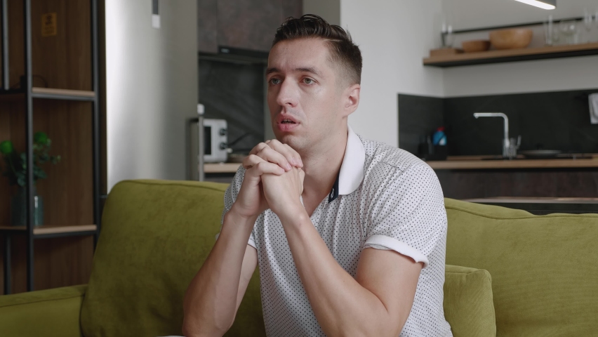 Young man asking for help about problem, praying to God. Religious male sit on sofa at home put hands in prayer. Guy praying with hope.