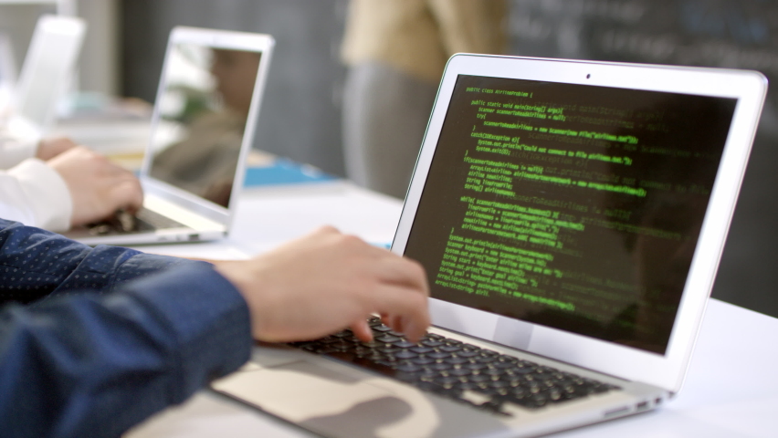 Closeup of laptop screen with program codes and male hands typing on keyboard while it teacher explaining material at blackboard