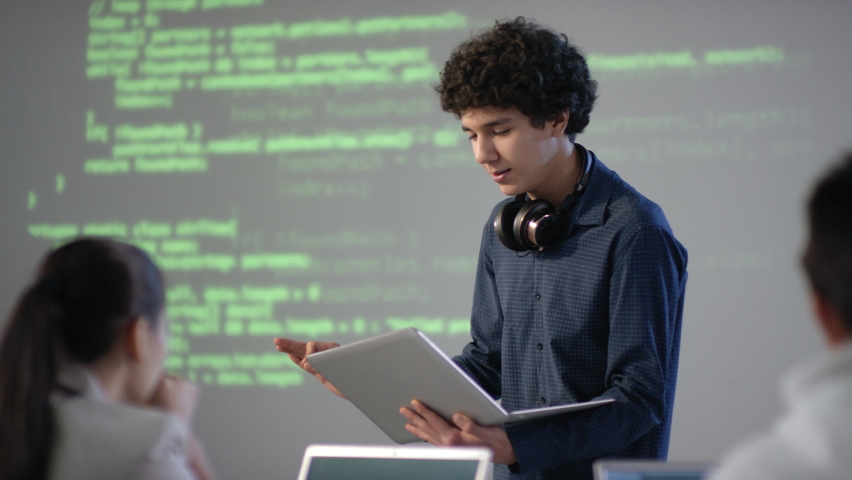 Medium closeup of cheerful mixed race student with laptop in hands making speech on computer programming topic smiling to classmates standing at board Royalty-Free Stock Footage #1061344393