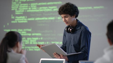 Medium closeup of cheerful mixed race student with laptop in hands making speech on computer programming topic smiling to classmates standing at board