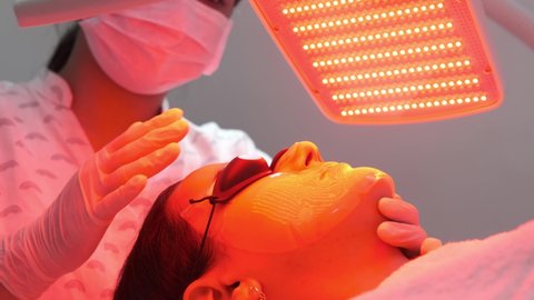 Young woman having red LED light facial therapy treatment in beauty salon. Hands of beautician adjusting collagen hydrogel mask on client face. Beauty, healthcare and wellness concept