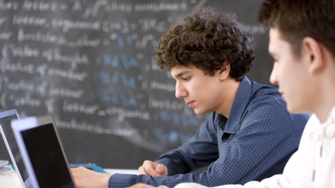 Medium closeup of two male multi-ethnic boys sitting at desks in modern classroom in front of laptops discussing subject of IT lesson with blackboard in background