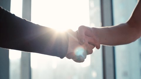 Close-up of two business partners shaking hands with each other to signify an agreement in the office against the background of the window and sunlight. Tracking shot in slow motion.