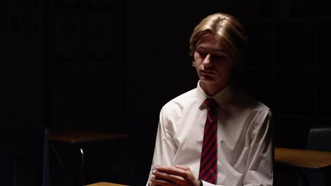 White teen boy student sitting at desk alone in dark classroom, depression