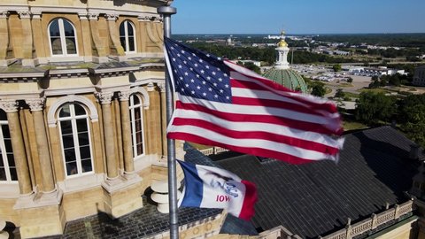 Drone aerial footage zooming out from Iowa State Capitol building with American flag and Iowa state flag waving in Des Moines on sunny day with view of skyline