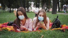 Students in medical masks during the Coronavirus pandemic using mobile apps holding smartphones looking at smartphones screen playing games texting messages enjoy social media applications in the park
