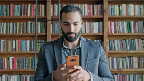 Young Man In The Library With Mobile Phone In His Hands. Guy With Beard And Black Hair. He Is Emotionally Playing Game On The Phone.