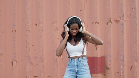 Young Black Girl Dancing On The Street Modern Dancing. She Is In Headphones. Girl Smiles And Enjoys Music And Dancing. Pretty Girl With Long Hair. She Is Wearing Jeans With Suspenders.