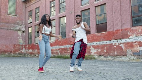 Street Dancing. Young Couple Dancing On The Street. Black Guy And Girl Move Rhythmically. They Make The Same Movements And Smile. Behind Them Are Old Houses. Cute Young People In Jeans. Stylish Guy
