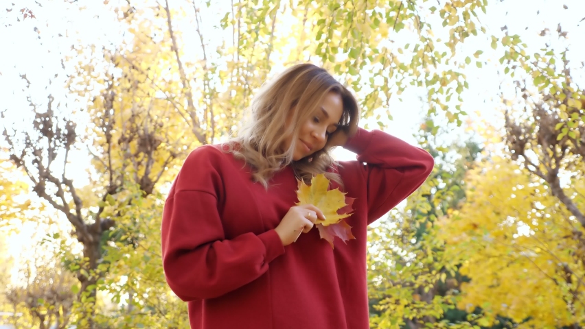 Positive young woman in red dress holds yellow maple leaves and looks at camera in autumn park