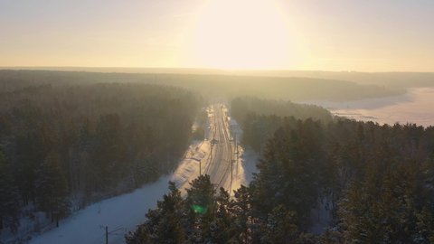 Aerial top-down view on sunrise over winter railway in pine forest under the snow.  Camera moves along railroad straight to the sun. Siberia, Russia.

