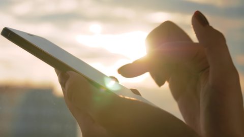 Close up side view: woman hand using smartphone device against warm sunset sky - scrolling and touching. Sun lens flare, golden hour. Relax, entertainment, leisure time and technology concept