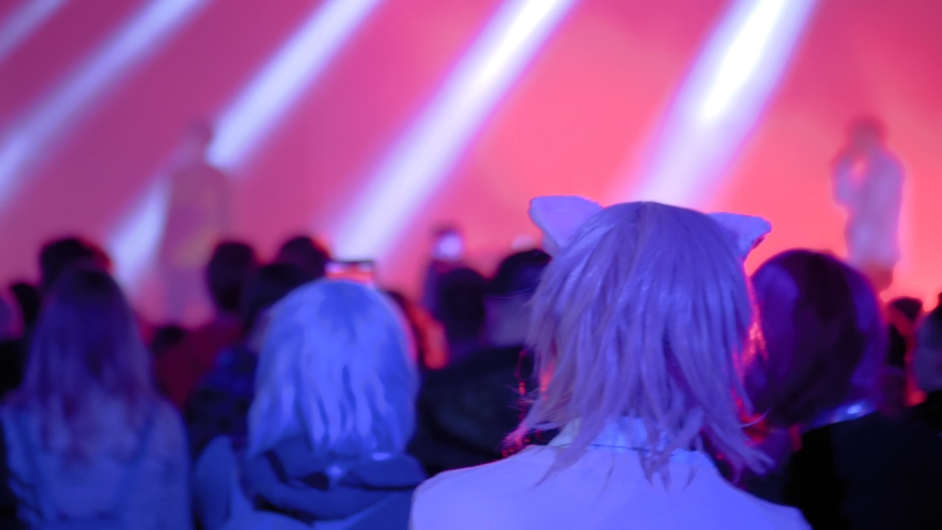 Slow motion: pink hair teenage girl with anime cat ear partying and cheering at rock concert in front of stage of nightclub. Bright colorful stage lighting. Nightlife and entertainment concept
